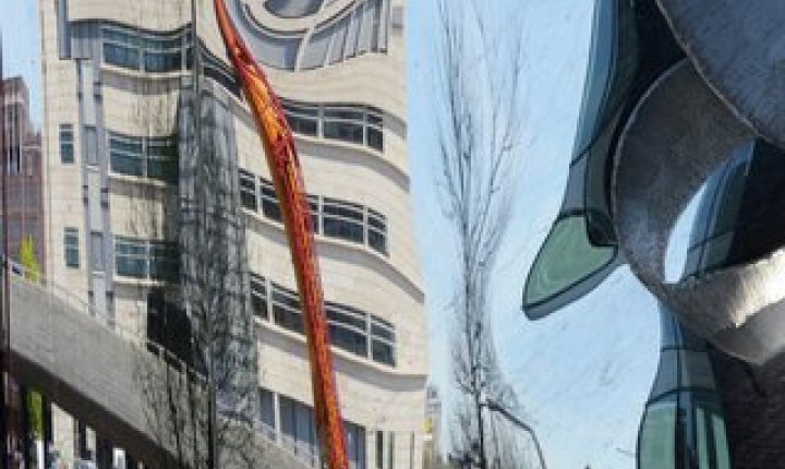 Urbant bybilde med høye skyskrapere, reflekterende glassfasader og en stor spiralformet metallskulptur i forgrunnen.