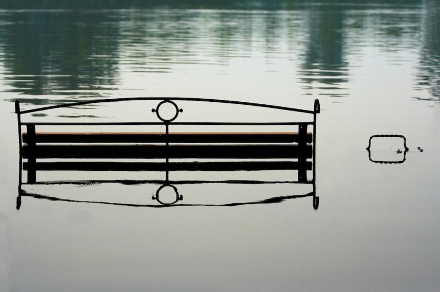 Parkbenk delvis nedsenket i rolig vann, noe som skaper en symmetrisk refleksjon mot en fredelig innsjøoverflate.